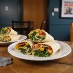 Close-up of The Vulture's Victory Veggie Wraps sliced on a counter in Brooklyn Nine-Nine precinct kitchen, displaying fresh vegetables and greens inside. The background features a Brooklyn Nine-Nine poster and badge coaster, emphasizing the show's identity and a casual lunch atmosphere.