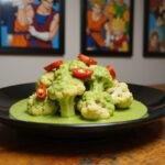 A realistic presentation of Piccolo's Special Beam Cannon Cauliflower featuring bright green cauliflower florets with red pepper accents, served on a black plate in an anime-themed kitchen with Dragon Ball Z posters behind, capturing the energetic vibe of the show.