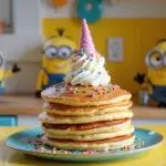 A realistic image of Agnes's fluffy unicorn pancakes on a bright kitchen counter, decorated with rainbow sprinkles, whipped cream, and a cute unicorn topper, set in a cheerful Minions-themed kitchen with yellow and blue decor, creating a fun kids' breakfast scene.