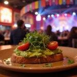 A close-up of a gourmet avocado toast with fresh avocado slices, microgreens, and cherry tomatoes, presented on a rustic plate amidst a lively circus-themed background featuring banners and bright lighting from The Greatest Showman movie scene.