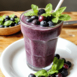 A vibrant blue blueberry smoothie in a clear glass, topped with fresh blueberries and a sprig of mint, set on a natural wooden table with scattered blueberries around, styled with a minimalist aesthetic and soft natural lighting.
