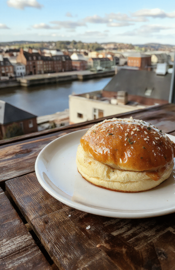 A vibrant, freshly assembled Belfast Baps sandwich depicted in bright natural light, featuring crispy bacon, black pudding, fresh greens, and a soft bun, styled on a rustic wooden table with a scenic Belfast cityscape in the background.