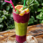 A vibrant green and purple layered tropical mocktail served in a tall glass with fresh fruit garnishes, colorful straws, and tropical leaves on a rustic wooden table, evoking an adventurous jungle scene with lush greenery in the background.