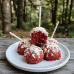 Colorful red velvet cake pops coated with glossy red icing, decorated with green candy leaves and tiny edible pearls, arranged on a white plate with a dark wooden background, styled to resemble poisoned apples from a fairy tale.
