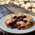 A vibrant slice of Bambi Forest Berry Pie displayed on a rustic wooden plate. The pie has a golden-brown crust with a glossy berry filling bursting with red and purple hues, topped with fresh mint leaves. The scene is set outdoors with soft sunlight highlighting the textures and colors, evoking a whimsical woodland atmosphere with scattered berries around.