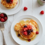 Colorful Mickey Mouse shaped pancakes stacked on a white plate, topped with fresh strawberries, blueberries, and a drizzle of syrup. The pancakes have a fluffy texture and golden-brown exterior, garnished with playful cartoon charm. Bright morning lighting enhances the vibrant colors, set on a cheerful breakfast table with a Disney-themed backdrop.