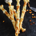 A spooky platter of Jack Skellington Skeleton Bone Breadsticks arranged on a dark slate board. The breadsticks are shaped like bones, with some decorated as skulls using black sesame seeds and olive slices for eyes. The scene features a dim, eerie lighting with Halloween-themed decorations in the background, creating a haunting, festive atmosphere.