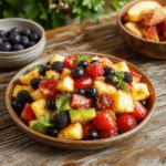 Colorful fruit salad arranged beautifully in a clear glass bowl featuring vibrant strawberries, kiwi slices, blueberries, orange segments, and grapes, glistening with juice, styled with mint leaves on top for a fresh look, set on a rustic wooden table with soft natural lighting