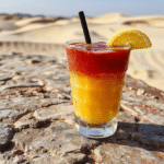 A vibrant, glowing Tatooine Sunset Punch served in a clear glass with ice cubes, garnished with tropical fruit slices and a mini umbrella. The punch's fiery orange and red hues mimic a sunset over a desert landscape, with a backdrop of a starry night sky and a sandy surface resembling Tatooine's dunes.
