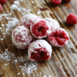 Colorful raspberry snowballs arranged on a white plate with powdered sugar dusted on top, showing soft, crumbly texture with vibrant red raspberry centers and delicate coating, styled on a rustic wooden surface with a festive holiday backdrop.