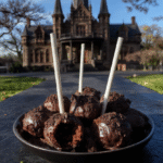 Bright black and gray Raven Cake Pops arranged on a dark, textured surface. The cake pops are decorated with intricate feather and raven details, with glossy finishes and edible metallic accents. Some pops are mounted on black sticks, with a subtle shimmer backdrop, evoking a mysterious Gothic aesthetic, perfect for a spooky celebration.