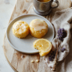 Two golden-brown lemon cakes with delicate lemon glaze drizzle, dusted with powdered sugar, placed on a rustic wooden cutting board beside a teacup and a sprig of lemon verbena, soft natural light, cozy and elegant presentation, inspired by Sansa Stark’s genteel style