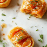 Elegant smoked salmon and dill pinwheels arranged on a white porcelain plate, each wheel sliced to reveal vibrant pink salmon, fresh green dill, and pale cream cheese layers, garnished with lemon zest and capers, against a soft blue linen background.