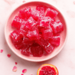 Vibrant neon-pink dragonfruit gummy bears arranged in a glossy pink plastic mold tray against a soft pastel pink background, with glittering sugar crystals and fresh dragonfruit slices nearby, sunlight casting soft highlights