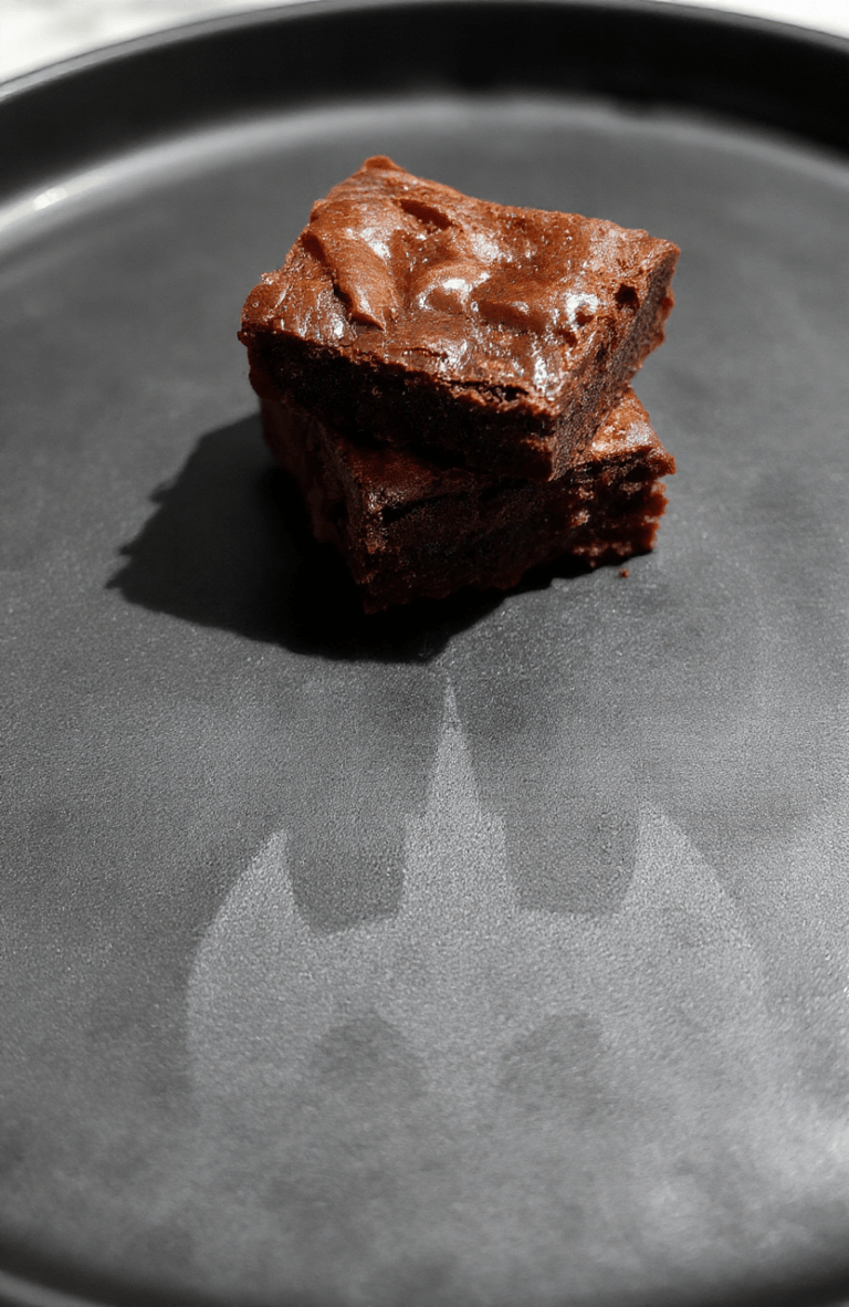 Rectangular dark chocolate brownies with a glossy cracked top, drizzled with white chocolate bat shapes and sprinkled with espresso granules, placed on a matte black ceramic plate with subtle gothic architectural engravings, soft shadows and natural window light casting a moody Gotham-inspired glow.