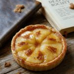 A golden-brown apple tart arranged in a rustic lattice pattern on a wooden board, surrounded by scattered autumn leaves and an open vintage book with