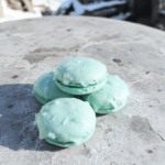 A pair of delicate, shimmering Elsa-themed macarons on a frosty white marble surface, with pastelblue and silver piped shells, edible glitter dusting, and tiny sugar snowflakes on top; soft background suggests a winter castle vibe with subtle pale blue and white tones.