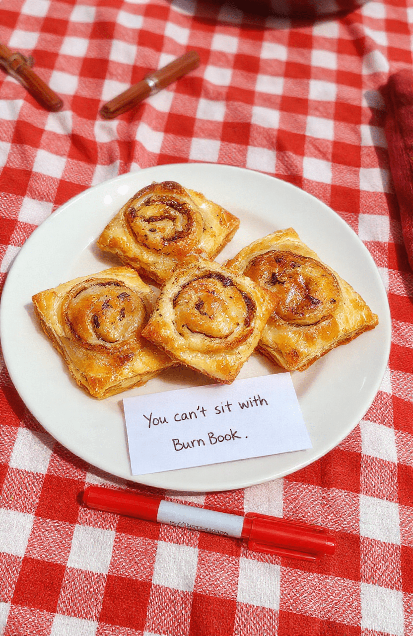 Golden-brown toaster strudel pastries with flaky layers, filled with vibrant raspberry and cream cheese swirls, dusted with powdered sugar and placed on a checkered red-and-white tablecloth. A small 'Burn Book' notebook and red marker peek out beside them, evoking the iconic Mean Girls aesthetic.