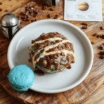 A vibrant, textured bark Candy Cloud with deep blue Swiss meringue buttercream, German chocolate shavings, and tiny edible gold leaf flecks arranged on a rustic wooden board, topped withminiature fondant Grogu ears and a dusting of edible silver glitter, under warm soft lighting with natural daylight highlights.