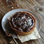 A sophisticated dark chocolate and sea salt tart on a rustic wooden board, with a glossy, deep brown chocolate ganache top, flaky buttery crust, and a delicate sprinkle of flaky sea salt crystals; a few broken shard pieces of dark chocolate and edible gold flakes accent the edge, set against a moody, dimly lit background with subtle parchment texture and a vintage copper whisk resting nearby.